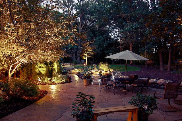 Landscape lighting illuminating an outdoor seating area that is paved with stone pavers. 