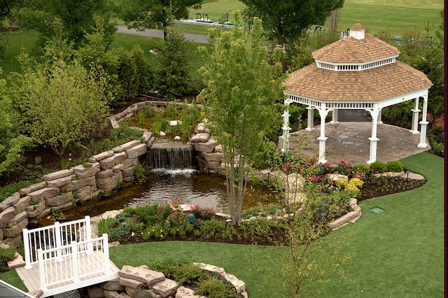 A landscape pond made out of stone in a grassy backyard adjacent to a white gazebo.  
