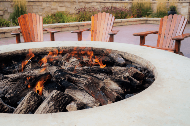  A close up of an outdoor gas fire pit surrounded by wooden Adirondack chairs  
