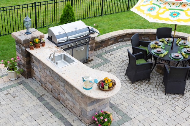 An outdoor kitchen with pavers, an outdoor gill, a bar with a sink, and a round table with chairs and an umbrella.