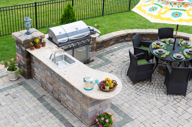 An outdoor kitchen with pavers, an outdoor gill, a bar with a sink, and a round table with chairs and an umbrella. 
