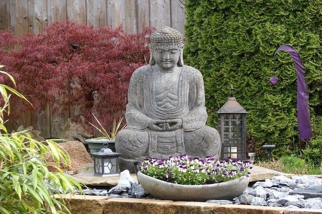 A grey stone statue of Buddha sitting in a rock garden with a Japanese maple tree in the background and other plants nearby.  Japanese garden and zen garden landscaping ideas.